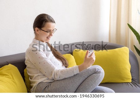 young cute caucasian woman communicates via video using a smartphone on yellow gray sofa for videocall meeting. Work online remote. Female entrepreneur working at home office confirming order on phone
