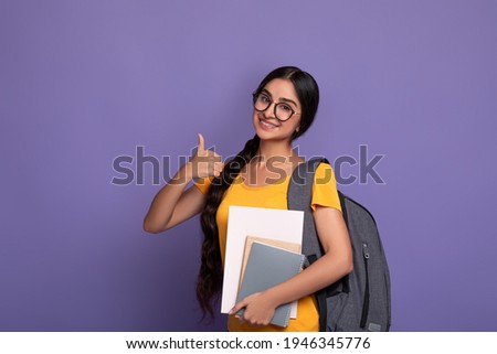 Education Concept. Portrait of smiling indian female student wearing eyeglasses showing thumbs up sign gesture, holding notebooks looking at camera. Woman wearing backpack, purple studio background Royalty-Free Stock Photo #1946345776