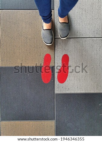 A woman waits outside the office with a pair of footmarks (red) on a sidewalk in the sun waiting for a financial transaction or shopping. Maintaining a social distance during the COVID-19 epidemic