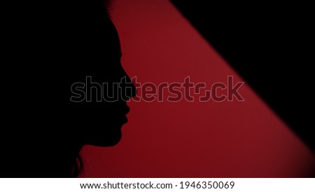 Side view of female face placing her finger on lips on dark red background. Closeup silhouette of serious woman showing hush gesture. Faded hand making keeping secret sign.