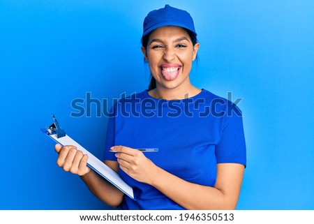Beautiful brunette woman wearing courier uniform holding clipboard sticking tongue out happy with funny expression. 
