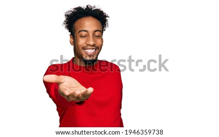 Young african american man with beard wearing casual winter sweater smiling cheerful offering palm hand giving assistance and acceptance. 