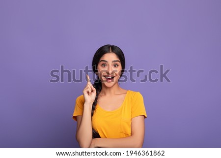 Inspiration, Creativity, Solution, Eureka concept. Portrait of excited young indian woman in yellow t-shirt raising finger up and smiling, having wow creative idea, purple studio background, copyspace Royalty-Free Stock Photo #1946361862