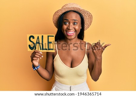Young african woman holding sale poster pointing thumb up to the side smiling happy with open mouth 