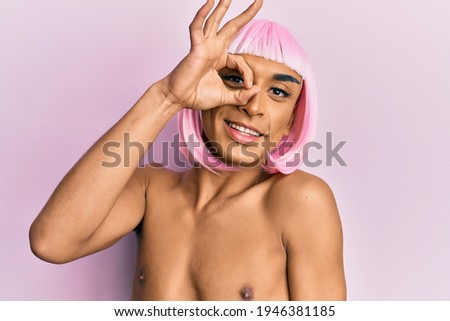 Hispanic transgender man wearing make up and pink wig smiling happy doing ok sign with hand on eye looking through fingers 