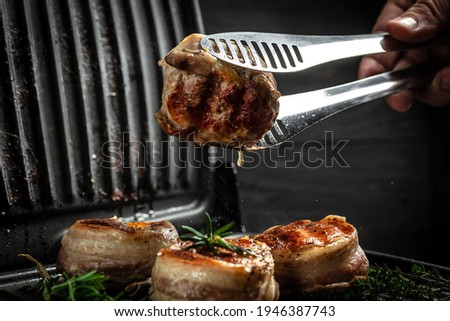 Medallions steaks from the beef tenderloin covered bacon on grill Dark background. Cooking beef steak on grill by chef hands.