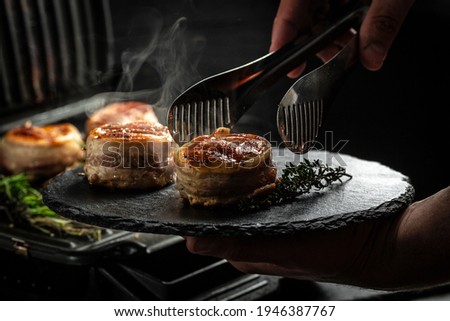 Medallions steaks from the beef tenderloin covered bacon on grill Dark background. Cooking beef steak on grill by chef hands.