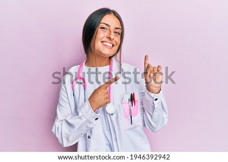 Beautiful hispanic woman wearing doctor uniform and stethoscope smiling and looking at the camera pointing with two hands and fingers to the side. 