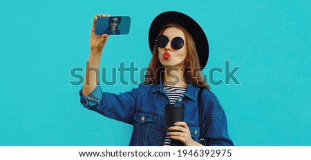 Portrait close up of stylish woman taking selfie picture by smartphone wearing a black hat, backpack on a blue background