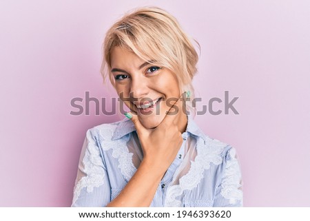 Young blonde girl wearing casual clothes looking confident at the camera smiling with crossed arms and hand raised on chin. thinking positive. 