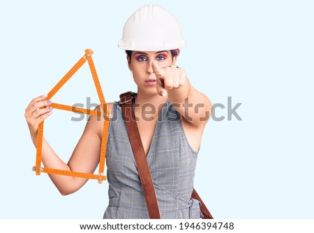 Young beautiful woman wearing architect hardhat holding build project pointing with finger to the camera and to you, confident gesture looking serious 