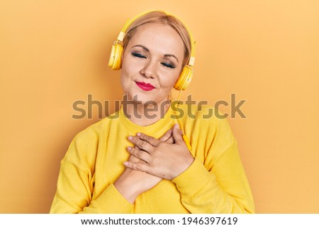 Young blonde woman listening to music using headphones smiling with hands on chest with closed eyes and grateful gesture on face. health concept. 