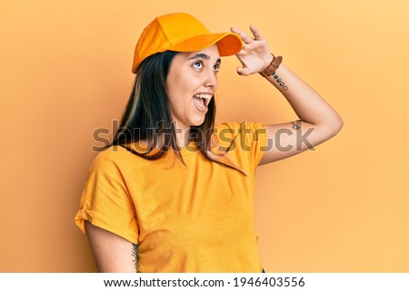 Young hispanic woman wearing delivery uniform and cap very happy and smiling looking far away with hand over head. searching concept. 