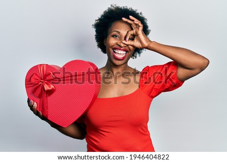 Young african american woman holding valentine gift smiling happy doing ok sign with hand on eye looking through fingers 