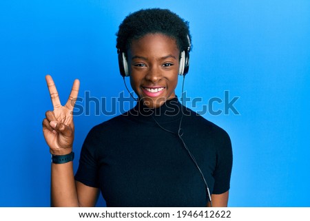 Young african american girl listening to music using headphones smiling with happy face winking at the camera doing victory sign. number two. 