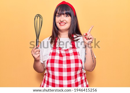 Young plus size woman wearing apron holding whisk smiling happy pointing with hand and finger to the side 
