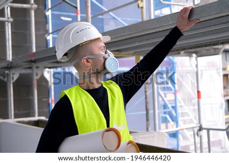 young male builder engineer, worker or architect in white hardhat and protective respirator during inspection in construction site, concept of monitoring repair of buildings, structures Royalty-Free Stock Photo #1946446420