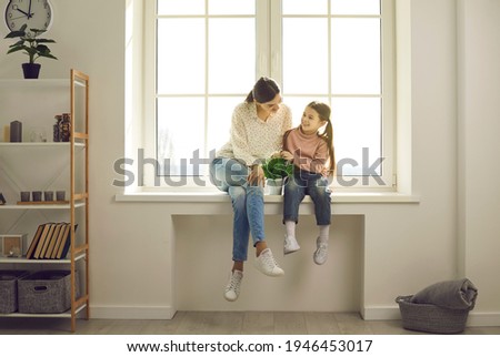 It's so cozy inside, let's stay at home. Happy family backlit by warm sunlight having fun in new house. Positive single mother together with little daughter sitting in sun on white plastic window sill Royalty-Free Stock Photo #1946453017