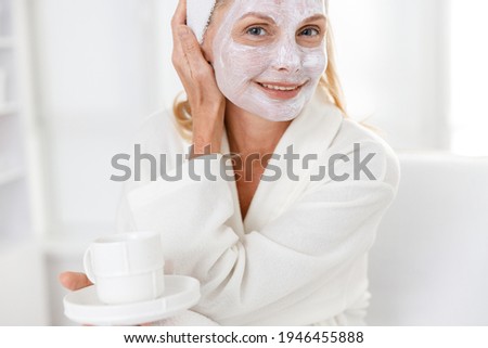Patient waits results of skin care treatment while drinking tea. Effective procedure. Senior woman in white bathrobe with mask on face.