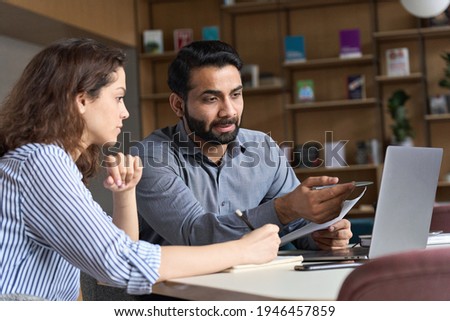 Professional indian teacher, executive or mentor helping latin student, new employee, teaching intern, explaining online job using laptop computer, talking, having teamwork discussion in office. Royalty-Free Stock Photo #1946457859