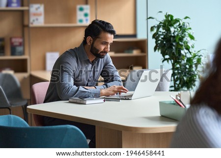 Young serious indian professional business man, eastern male student working on laptop, remote studying, distance learning, browsing using computer sitting at table in modern coworking office space. Royalty-Free Stock Photo #1946458441