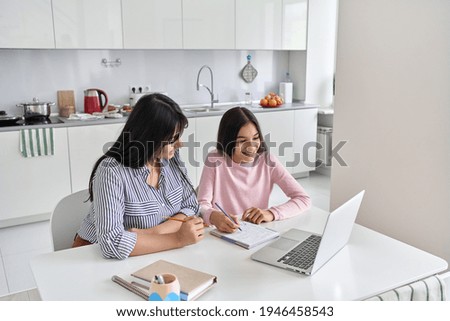 Happy young indian parent mother helping teenage child daughter remote learning online virtual class on laptop together at home. Teen school kid girl studying in kitchen with mum sit at kitchen table. Royalty-Free Stock Photo #1946458543