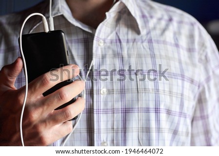 Caucasian young adult man wearing striped shirt holding mobile phone with earphones connected, blurry background.