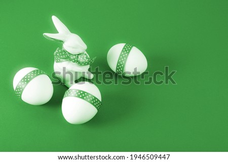 Easter Concept. Porcelain rabbit with three white eggs decorated with green ribbon on green paper background. Copy space. 