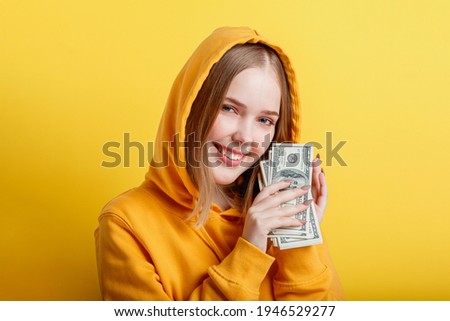 Emotional happy teenage blonde girl in hood win money cash holding dollars in hands isolated on color yellow background. Portrait young excited smiling woman with stack of money banknotes.