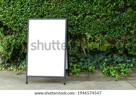 Blank white outdoor advertising stand or sandwich board mock up template. Clear street signage board placed outdoor by green tree hedges Background texture of standee at front of green tree wall.