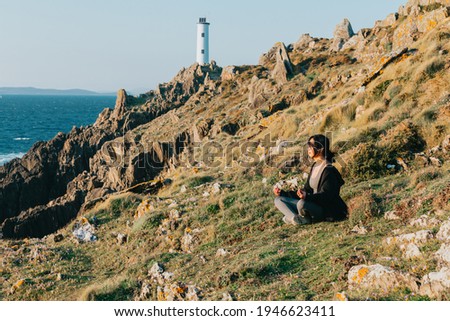 Young fit woman doing yoga in the rocks of a wild coast with a lighthouse as the background sunny day with copy space relax concept