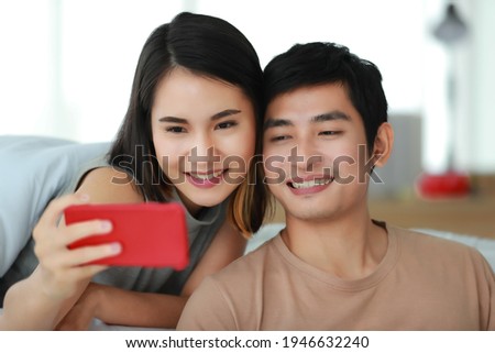 Portrait shot of Asian lover couple relaxes at home on the holidays. The wife lies on bed behind her husband that sits on the floor and embracing him. Woman playing social media on a smartphone
