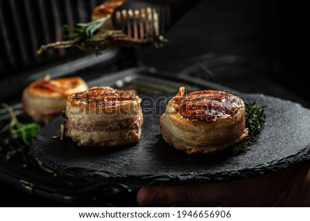 Medallions steaks from the beef tenderloin covered bacon on grill with smoke dark background. Cooking beef steak on grill by chef hands.