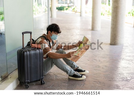 Travel, A man wearing glasses with a striped shirt and mask sit on the floor, reading map in his hand with suitcase placed on right side and backpack.