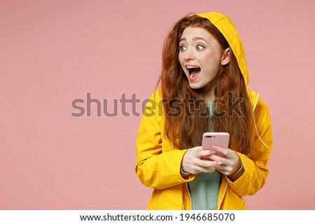 Redhead young woman in yellow waterproof raincoat hood outerwear hold mobile cell phone look aside isolated on pastel pink background studio Outdoors lifestyle wet fall cold weather season concept.