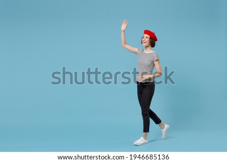 Full length of young smiling woman with short hairdo in french beret red hat striped t-shirt hold hands crossed folded walk going waving hand greet isolated on pastel blue background studio portrait