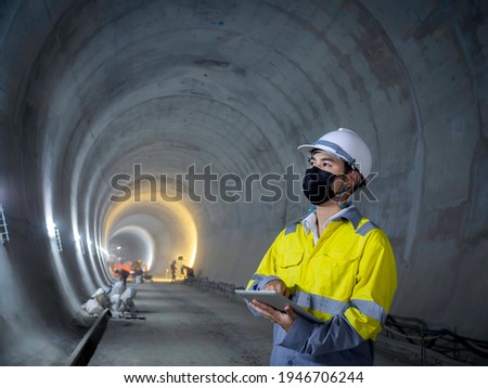 Young Asian tunnel engineering wearing high visibility jacket, face mask and white safety helmet working and using digital tablet in dark railway tunnel construction site area with copy space. Royalty-Free Stock Photo #1946706244