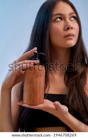 Organic cosmetic. Vertical shot of a young attractive asian woman holding cosmetic product and looking upside while posing against blue background. Beauty and wellness concept