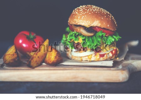 Fresh craft beef burger with vegetables, mushroom, lettuce, tomatoes, cheese and sauces on a wooden rustic board on a black background. Delicious grilled home made hamburger. Fast food background.