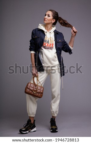 High fashion photo of a beautiful elegant young woman in a pretty white sports suit with hood, denim jeans jacket, sneakers, walking style, handbag posing over gray background. Studio Shot. Portrait.
