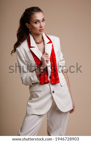 High fashion photo of a beautiful elegant young woman in a pretty white suit, jacket, trousers, pants, shoes, red accessories posing over beige powdery background. Studio Shot. Portrait. Slim Figure