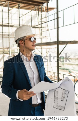 Young man architect wearing formal suit and hard hat during building construction control holding a blueprints on a construction site