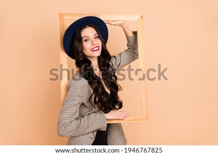 Photo of cute adorable young woman wear plaid jacket headwear inside wooden frame isolated beige color background