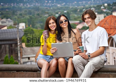 Friends sitting on the curb with a laptop. Teenagers with disposable paper cups drinking coffee against the backdrop of the city. Online learning concept.