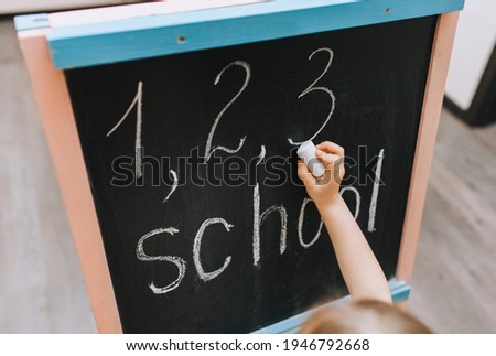The girl writes beautifully, diligently, draws on a black wooden board, easel, holding white chalk, the word school and numbers in her hand and fingers. Photography, copy space, homework.
