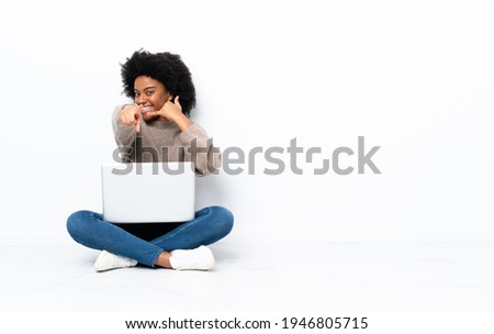 Young African American woman with a laptop sitting on the floor making phone gesture and pointing front
