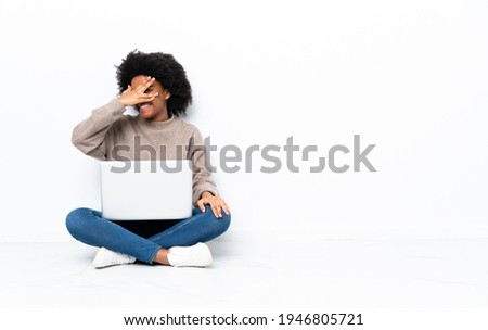 Young African American woman with a laptop sitting on the floor covering eyes by hands and smiling