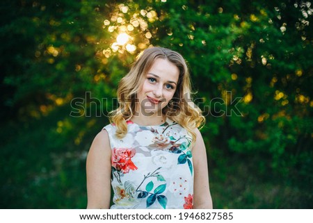 Beautiful blonde girl in a floral dress at sunset. Sunny portrait of a happy woman. Blonde on a green background. Portrait of a woman in the summer park