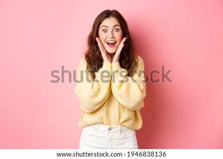 Excited beautiful girl staring surprised and amazed, holding hands on cheeks and gasping, looking with astonished face at promotional offer, standing near copy space pink background Royalty-Free Stock Photo #1946838136