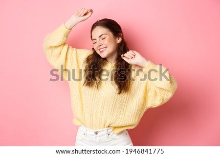 Portrait of beautiful caucasian woman having fun, dancing and enjoying party, raising hands up in relaxed, carefree pose, standing against pink background